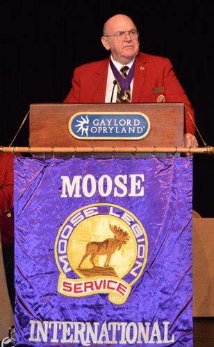 A man in red jacket standing at podium with moose legion banner.